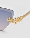 Close-up of For Art's Sake® Florist sunglasses, showcasing gold metal frames with sculpted temple accents and floral details. The blue gradient lens fades to a lighter hue, all set against a plain light gray background.