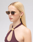 A woman with straight blonde hair wears For Art's Sake® Florist narrow, dark frameless sunglasses, gold earrings, and a purple halter top with a gold clasp. She faces slightly sideways against a plain light background.
