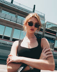 A woman in For Art's Sake® Florist floral sunglasses and a black sleeveless top stands with arms crossed before a modern racing stadium with glass windows and checkered details.