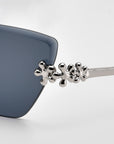 Close-up of For Art's Sake® Florist sunglasses with dark blue lenses and a silver metallic frame. Sculpted floral accents on the arm near the lens add an elegant decorative touch.