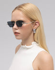 A woman with long blonde hair wears For Art's Sake® Florist black floral sunglasses, a sleeveless white ruffled-collar top, and square silver earrings against a plain light background.