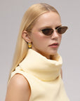 A woman with straight blond hair wears For Art's Sake® Lake cat-eye sunglasses with sculpted temples, gold earrings, and a sleeveless high-neck cream top, gazing slightly to the side against a neutral background.