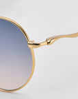 Close-up of For Art's Sake® Lines: oversized aviator sunglasses with a gold frame and gradient blue-to-clear lenses. Sculpted temples and hinge detail stand out against a plain light gray background, showcasing the brand’s signature style.