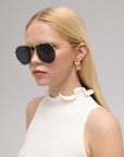 A woman with long blonde hair wears For Art’s Sake® Lines oversized aviator sunglasses with sculpted temples, gold earrings, and a sleeveless white top with a ruffled collar, posing against a neutral background.