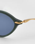 Close-up of For Art's Sake® Totto sunglasses featuring a green acetate and metal frame, blue-tinted lenses, and gold sculpted temples on a plain light gray background.