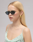 A woman with long blond hair wears For Art's Sake® Totto sunglasses with sculpted temples, gold rectangular earrings, and a light green polka-dot top with thin straps. She faces slightly to the side against a neutral background.