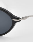 Close-up of the hinge and frame corner of For Art's Sake® Totto sunglasses with sleek acetate and metal construction, dark lenses, and a silver arm against a light gray background.