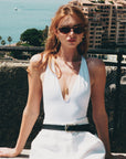 A woman with long red hair, wearing For Art's Sake® Totto sunglasses with sculpted acetate and metal temples, a white sleeveless top, and white pants with a black belt, sits outside in sunlight, city buildings and the sea behind her.