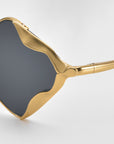 Close-up of For Art's Sake® Leaf I sculptural eyewear, showcasing gold, wavy-edged frames and dark tinted lenses in a statement design against a light background.
