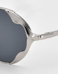 A close-up of For Art's Sake® Leaf II sculptural sunglasses featuring dark lenses and a shiny, wavy silver metal frame, displayed against a plain white background.