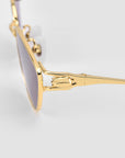 Close-up of For Art's Sake® Tulip II glasses with gold sculptural frames, purple-tinted lenses, intricate hinge detail, and a smooth reflective metal finish on a light gray background.