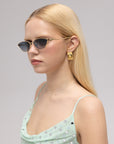 A woman with straight blonde hair wears For Art's Sake® Tulip II black cat-eye sunglasses with a bold acetate front, large gold earrings, and a light green polka-dot dress, gazing to the side against a neutral background.