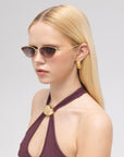 A woman with straight blonde hair wears For Art's Sake® Tulip II bold acetate sunglasses, gold geometric earrings, and a dark halter top with a gold accent, posing against a plain light background.