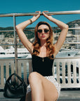 A woman in For Art's Sake® Tulip II sunglasses, a black top, and polka dot skirt sits with one leg crossed and arms raised on a rail. City buildings, mountains, yachts, and a black handbag complete the sunny backdrop.