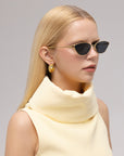 A woman with straight blonde hair wears For Art's Sake® Tulip II bold acetate cat-eye sunglasses, gold geometric earrings, and a sleeveless pale yellow high-collar top against a neutral background.