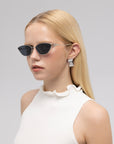 A woman with straight blonde hair models For Art's Sake® Tulip II bold acetate sunglasses, paired with rectangular silver earrings and a sleeveless white ruffled top, posing before a light grey backdrop.