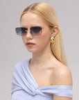 A woman with long blonde hair wears For Art's Sake® Magnolia frameless sunglasses, gold square earrings, and a light blue off-shoulder top, standing against a neutral background.