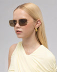 A young woman with straight blonde hair wears For Art's Sake® Magnolia sculptural sunglasses, gold geometric earrings, and a cream draped sleeveless top, looking slightly to the side against a plain background.