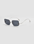 The Magnolia by For Art's Sake® are rectangular, frameless sunglasses with dark lenses and slim, curved metal arms, shown on a plain light gray background.