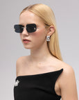 A woman with straight blonde hair poses against a plain light background, wearing For Art's Sake® Magnolia frameless sunglasses, rectangular earrings, and a black strapless dress with a jeweled brooch.