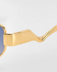 Close-up of Abstraction II by For Art's Sake®: statement eyewear with shiny gold geometric metal frames, wavy arms, and blue-tinted lenses, set against a plain light background.