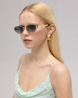 A woman with straight blonde hair wears For Art's Sake® Abstraction II geometric metal sunglasses, gold rectangular earrings, and a light green sequin top, gazing slightly to the side against a plain background.