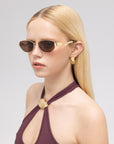 A woman with straight blonde hair wears For Art's Sake® Abstraction II geometric metal sunglasses, gold earrings, and a sleeveless maroon top with a gold clasp, gazing to the side against a plain background.