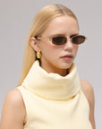 A woman with straight blonde hair wears For Art's Sake® Mesa I narrow sculptural sunglasses, a sleeveless high-neck cream top, and gold geometric earrings, posing against a plain light background.