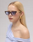 A woman with long, straight blonde hair wears For Art's Sake® Mesa II angular luxury sunglasses, gold earrings, and a light blue off-shoulder top while looking to the side against a neutral background.