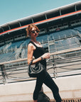 A woman in For Art's Sake® Mesa I sculptural eyewear, a black tank top, and leggings walks by empty stadium bleachers under a clear blue sky, carrying a black woven handbag.