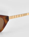 Close-up of For Art's Sake® Mesa II luxury cat-eye sunglasses featuring a brown acetate frame and gold textured arms, set against a plain light gray background.
