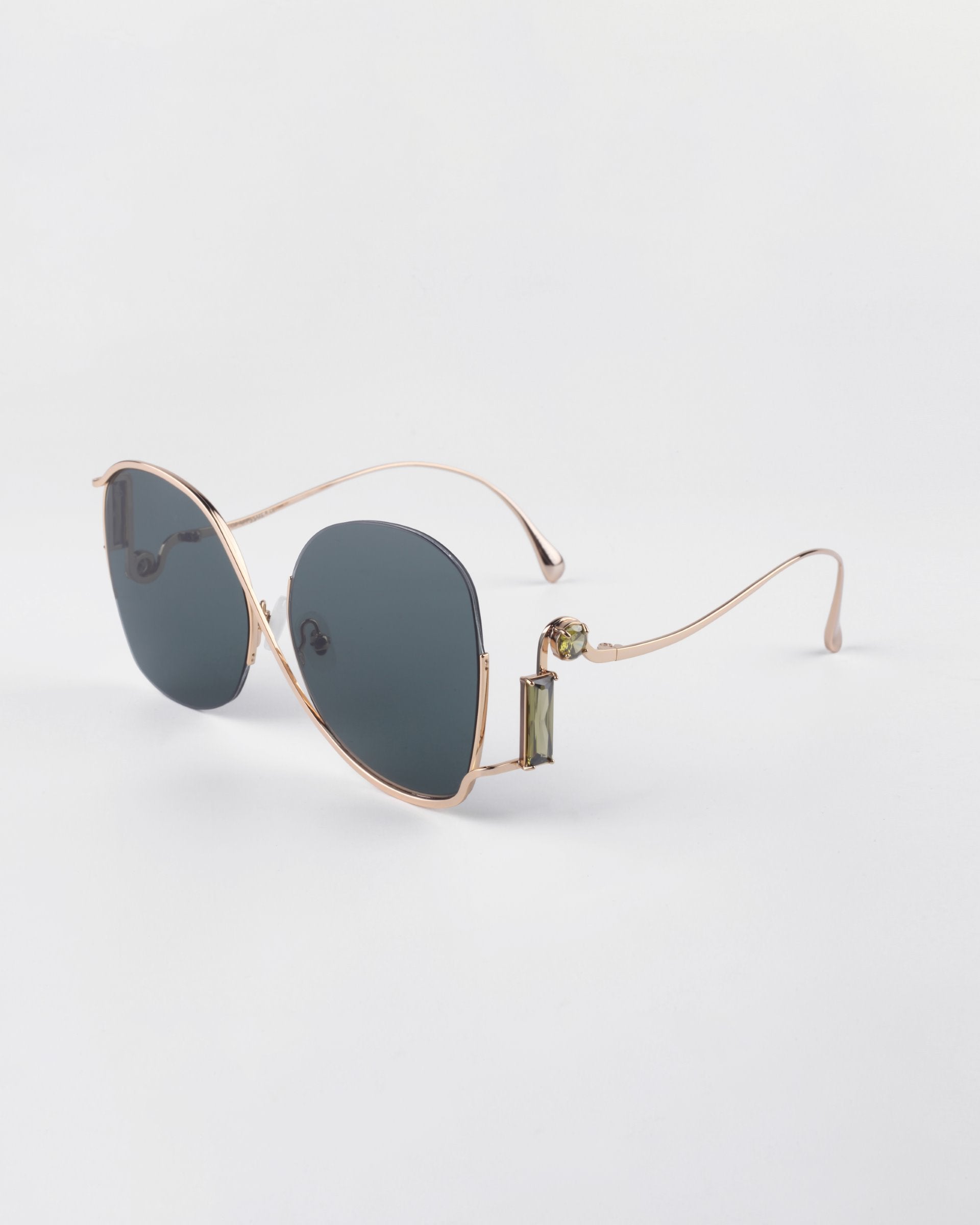 A pair of luxurious sunglasses featuring gold metal frames and dark, UV-protected lenses. The design includes round lenses and thin, curved temple arms with small green decorative details near the hinges. Crystal embellishments add a touch of elegance against a plain white background. These are the Sapphire sunglasses by For Art's Sake®.