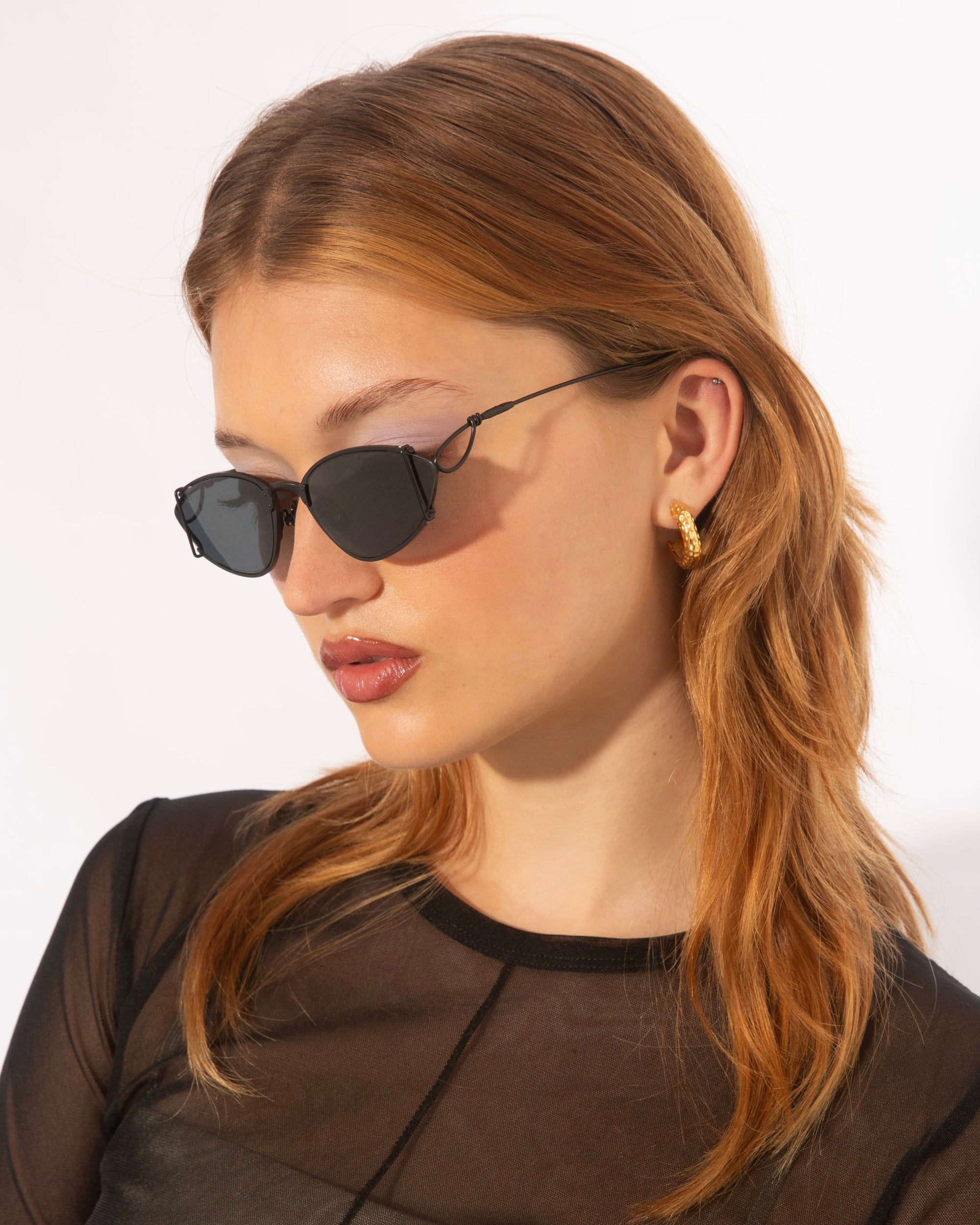 A person with light skin, long auburn hair, and wearing For Art's Sake® Ornate sunglasses with ultra-lightweight nylon lenses is shown in profile against a plain background. They are dressed in a sheer black top and gold hoop earrings that shimmer like 18-karat gold-plated jewelry.