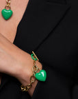 A person wearing a black blazer showcases The Kiss Bracelet by For Art's Sake®, an 18k gold chain bracelet adorned with an enamel-covered heart charm. A matching necklace with a green heart pendant is partially visible. The person's hand is tucked into the blazer.