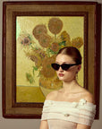A woman in For Art's Sake® Vincent Van Gogh limited edition cat-eye sunglasses and an off-shoulder white dress poses by a framed sunflower painting.