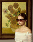 A woman wearing For Art's Sake® Vincent Van Gogh limited edition cat-eye sunglasses and an off-the-shoulder white top poses before a sunflower painting with a yellow background reminiscent of Van Gogh’s iconic style.