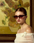 A woman in cat-eye sunglasses by For Art's Sake® and an off-shoulder white top poses before a framed Vincent Van Gogh painting.