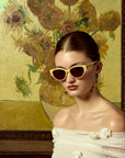 A woman in off-the-shoulder white attire and cream cat-eye sunglasses poses before a sunflower painting, her look completed with dangling earrings—showcasing the limited edition Vincent Van Gogh by For Art's Sake®.