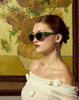 A woman in For Art's Sake® Vincent Van Gogh limited edition cat-eye sunglasses and a white off-shoulder top poses in profile before a framed sunflower painting, her hair tied back and wearing dangling earrings.