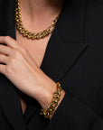 A close-up image of a person wearing a chunky gold chain necklace and the Wave Bracelet Gold by For Art's Sake®. The person is dressed in a black outfit, with their hand gently touching their chest, highlighting the jewelry against the dark fabric and evoking an elegant 80s jewellery vibe.