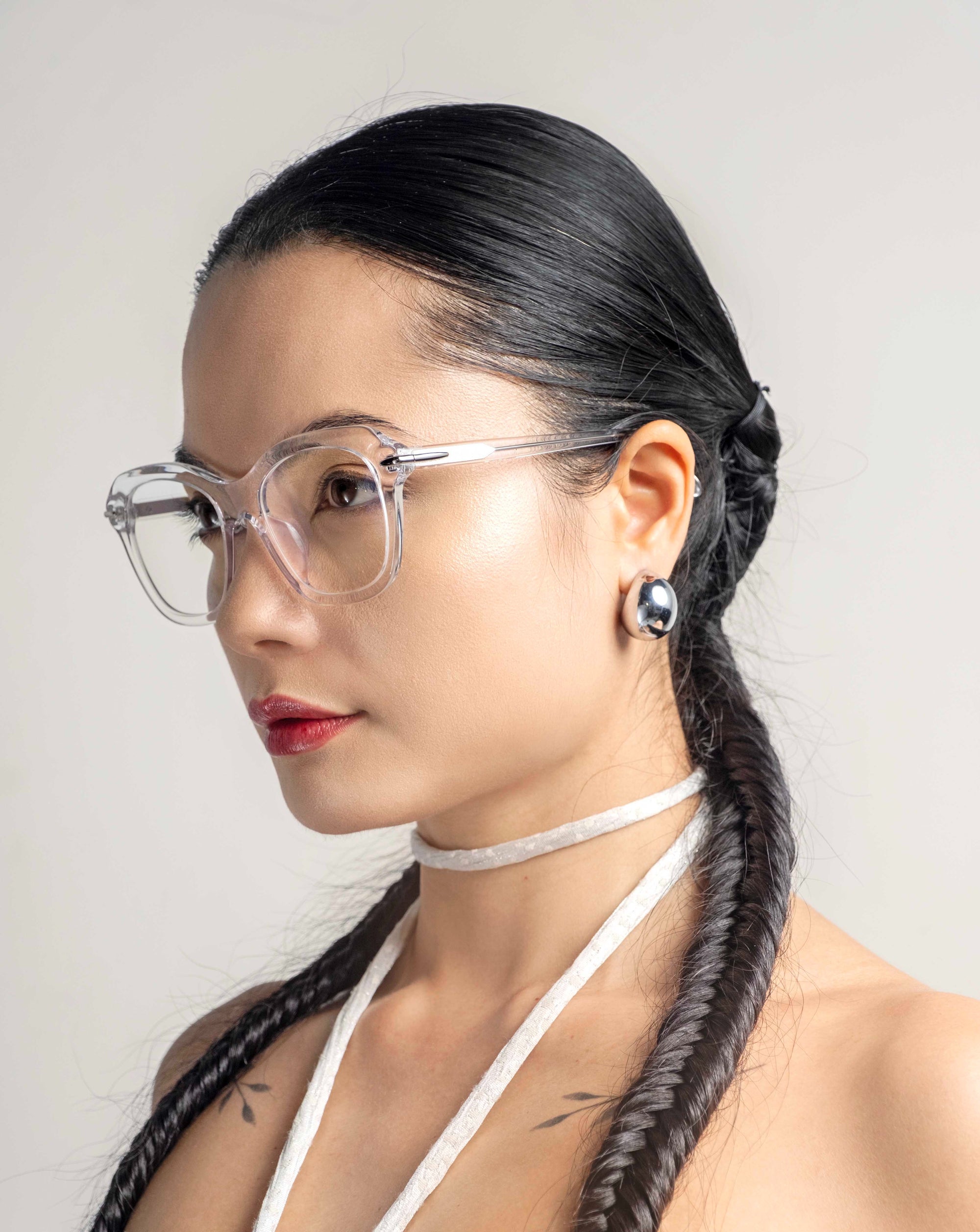 A person with long, dark braided hair, wearing clear-framed, cat-eye silhouette glasses and a white halter strap, is pictured in a profile view against a plain background. They have a calm expression with shiny Helene earrings by For Art's Sake® visible.