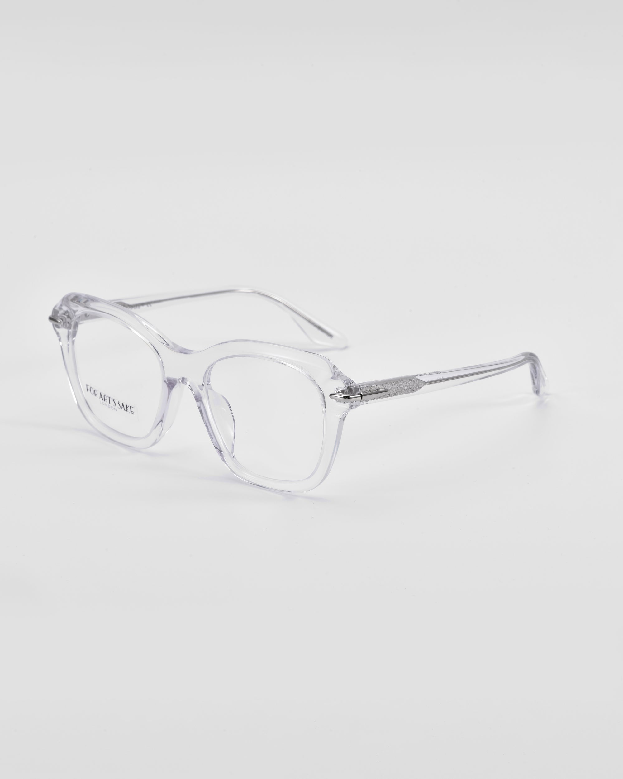 A pair of clear, transparent Helene by For Art's Sake® eyeglasses with square frames is placed on a white surface. The lenses are large with a slight retro style, and the temples feature 18-karat gold plating near the hinges. The glasses have a simple and minimalist design perfect for those seeking both elegance and functionality.