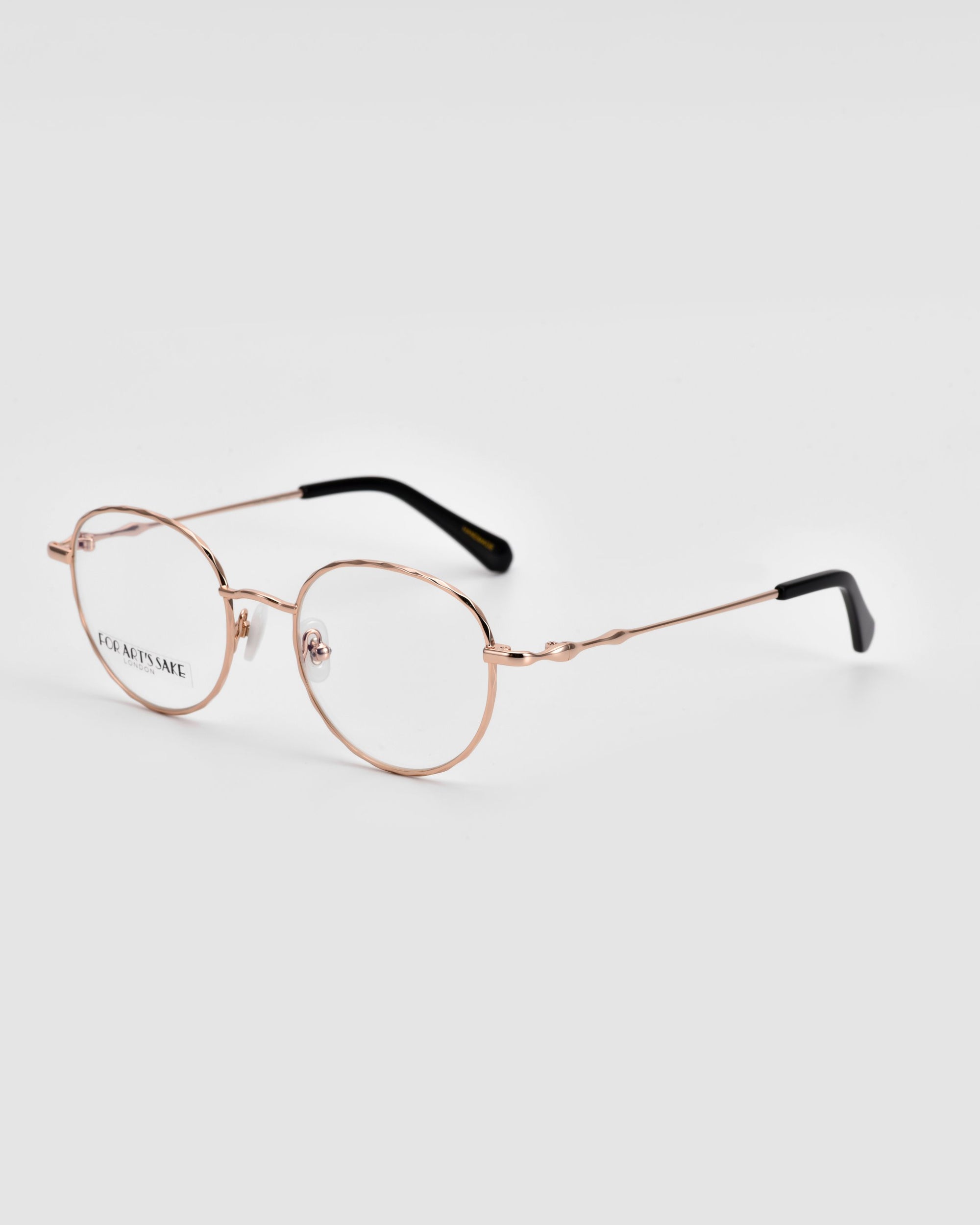 Delicate, gold-rimmed Bone I eyeglasses with oval lenses by For Art's Sake® feature "JOC AETSNAE" printed on one lens. The temples boast luxurious metal detailing and culminate in black tips. Set against a plain light grey background, these glasses exude sophistication.