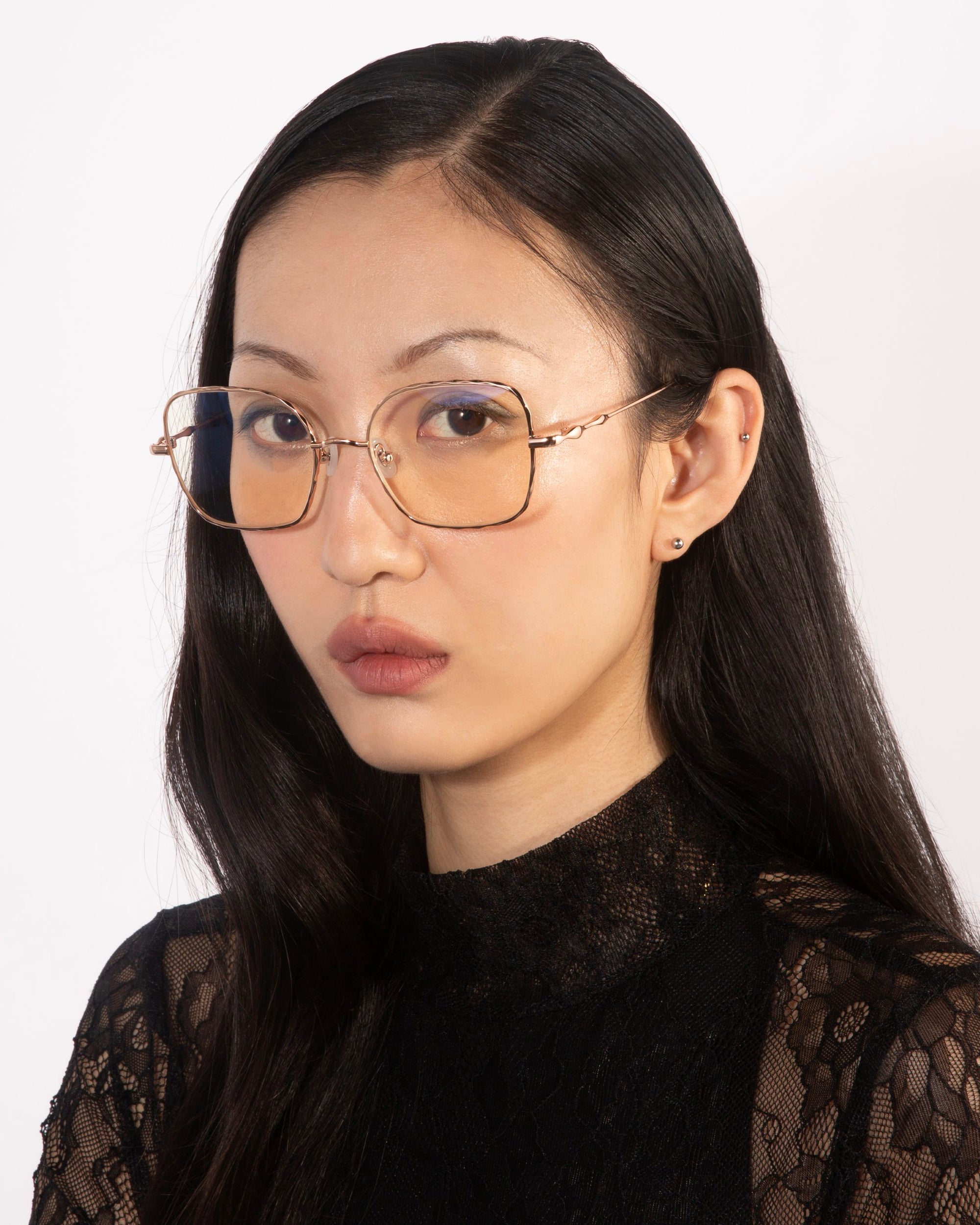 A woman with long black hair is wearing a black lace top and For Art's Sake® Bone I large square-framed glasses with a slight tint, featuring jade stone nose pads. She is looking slightly to the side with a serious expression. The background is light-colored and plain.