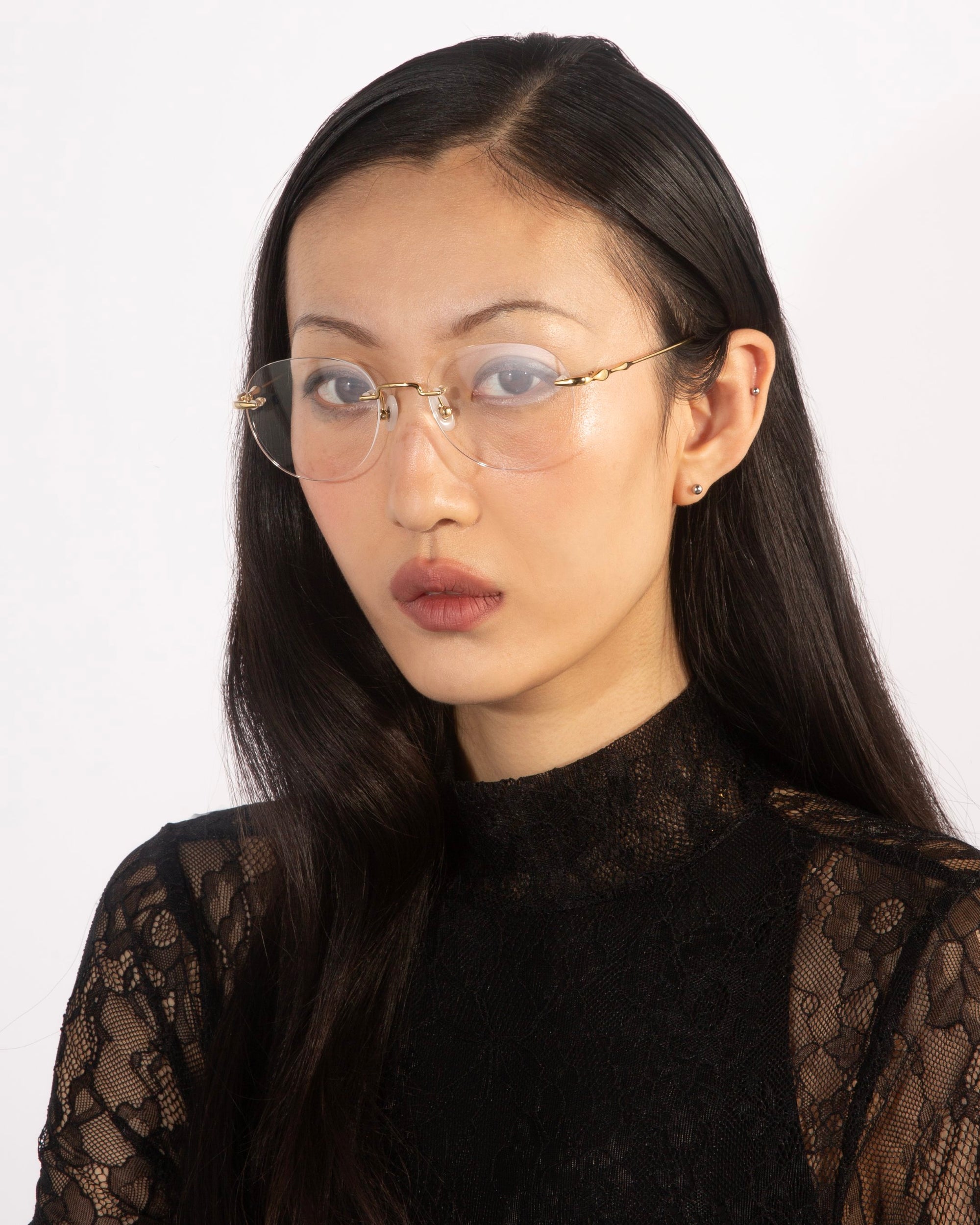 A person with long black hair and wearing glasses is looking directly at the camera with a neutral expression, exuding refined elegance in a "Misty" black lace top by For Art's Sake®. They stand against a plain white background, embodying contemporary design.