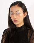 A person with long black hair and wearing glasses is looking directly at the camera with a neutral expression, exuding refined elegance in a "Misty" black lace top by For Art's Sake®. They stand against a plain white background, embodying contemporary design.