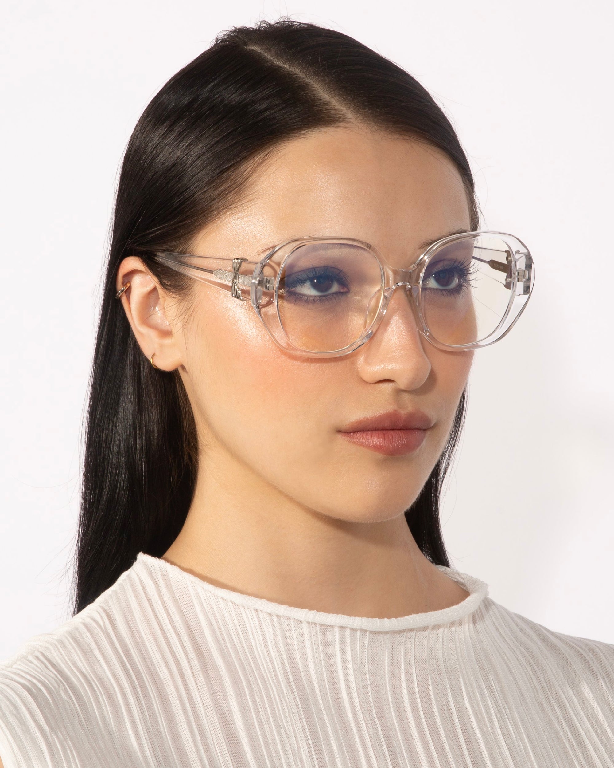 A person with long, straight black hair is wearing the For Art's Sake® Bow eyeglasses, which feature large, transparent lenses and a unique vintage design. The individual has a serious expression and is dressed in a white, textured high-neck top against a plain white background.