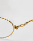 This close-up of the gold Thorn eyeglass frames by For Art's Sake® features a delicate rose detail on the hinge. The polished finish adds elegance, making it a true statement piece. The neutral background complements its exquisite oval silhouette beautifully.