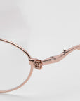 Close-up of For Art's Sake® Thorn eyeglasses, showcasing a delicate rose detail on the temple arm. The modern oval silhouette frame is in a soft rose gold color with a smooth, elegant finish against a neutral background.
