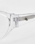 The Rosalia I by For Art's Sake® features transparent eyeglasses with a bold frame and cat-eye design, complemented by visible silver hinges and textured inner arms. Part of the Rose Garden Collection, these elegant glasses are pictured on a plain white background.